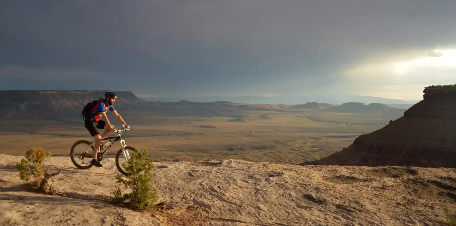 Mountain biker rolling the slickrock mesa top of Gooseberry under a dramatic stormy sky, the valley spread out far below