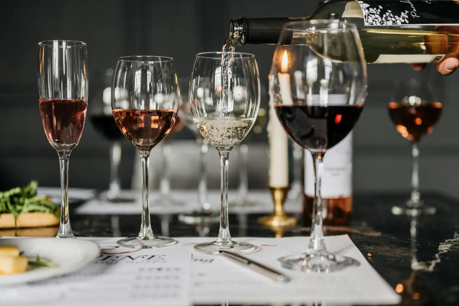A wine flight on a marble bar — rosé, sparkling, white and red pours with white being poured from a bottle