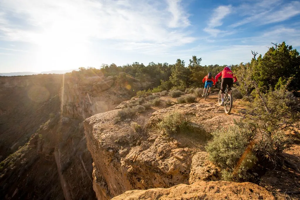 Two mountain bikers riding slickrock benches along a mesa rim at golden hour, the cliff dropping away to the valley below