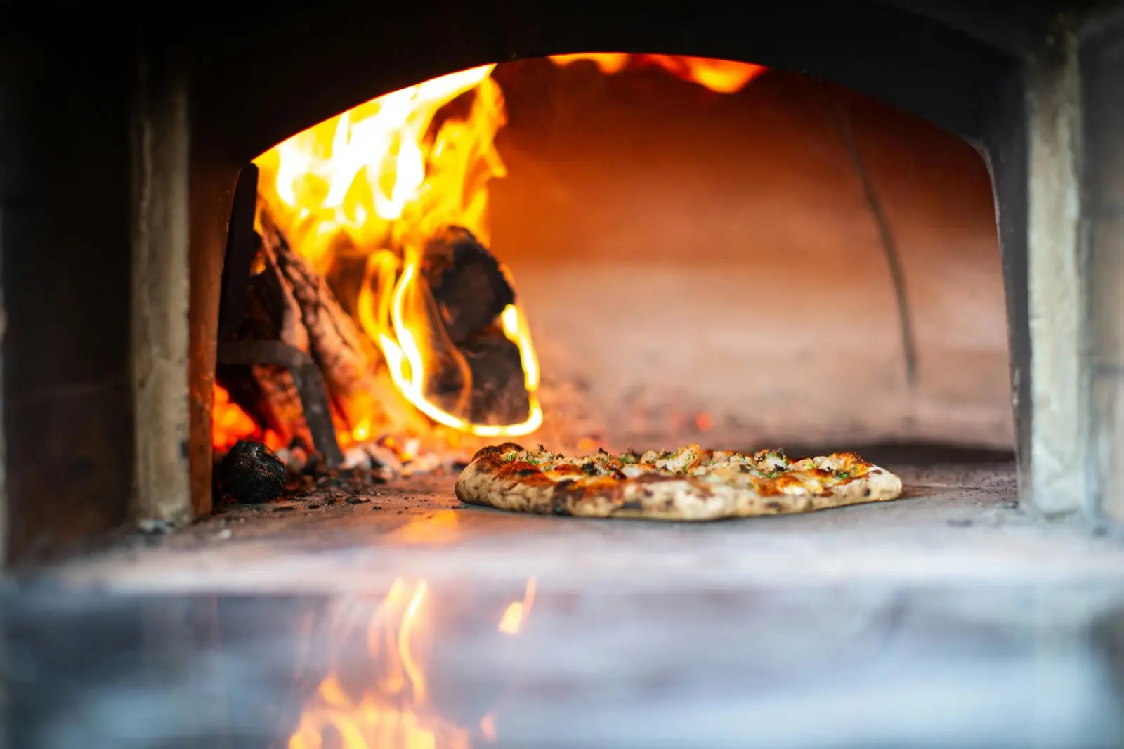 A wood-fired pizza emerging from a brick oven with flames glowing in the background