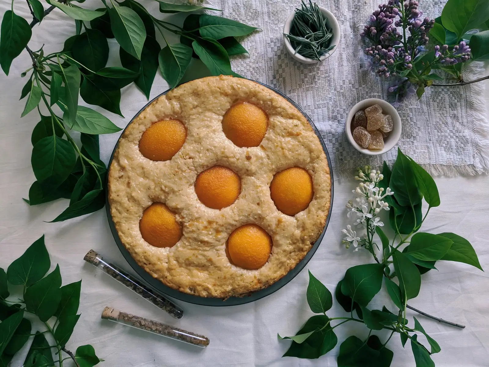 A homemade peach pie with golden peach halves arranged on top, surrounded by lilacs and citrus leaves