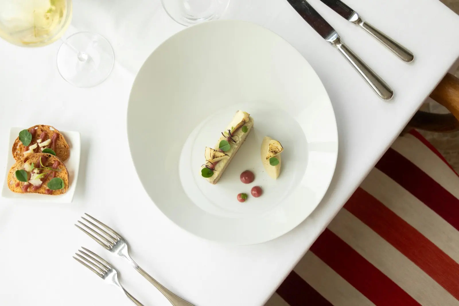 Elevated fine dining plate with composed garnish, crostini, and a glass of white wine on a linen-set table